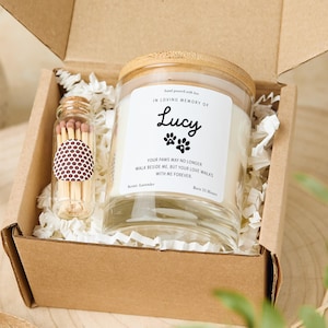 Puede incluir: Un juego de regalo en una caja de cartón que contiene una vela de tarro de vidrio con tapa de madera, un pequeño vial de vidrio de cerillas y relleno de papel blanco. La vela tiene una etiqueta que dice "IN LOVING MEMORY OF Lucy" con huellas de patas.