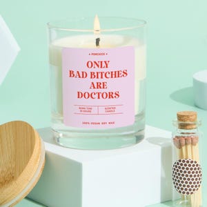 May include: A lit scented candle in a clear glass jar with a pink label that reads "ONLY BAD BITCHES ARE DOCTORS." The candle is made of 100% vegan soy wax and has a burn time of 25 hours. A small glass bottle of matches is next to the candle.