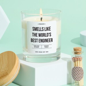 Pode incluir: Uma vela perfumada acesa num frasco de vidro transparente com o texto "Smells Like The World's Best Engineer". A vela tem um tempo de queima de 25 horas e é feita de cera de soja 100% vegana. Uma pequena garrafa de vidro com fósforos está ao lado.