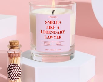 May include: A lit scented candle in a clear glass jar with a pink label that reads "Smells Like a Legendary Lawyer." A small glass bottle of matches and two loose matches are also in the image. The candle is made of 100% vegan soy wax.