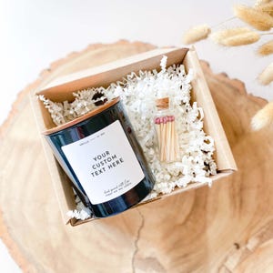 以下が含まれることがあります： 銅製の蓋と「Vanilla Soy Wax」と「Your Custom Text Here」と書かれた白いラベルが付いた黒いキャンドル。キャンドルは、白いシュレッダーペーパーと小さなガラス瓶のマッチが入った小さな茶色の箱に入っています。