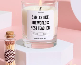 May include: A lit scented candle in a clear glass jar with the text "Smells Like The World's Best Teacher." A small glass bottle with a cork stopper and matches is next to the candle. The candle is on a white geometric stand against a pink background.