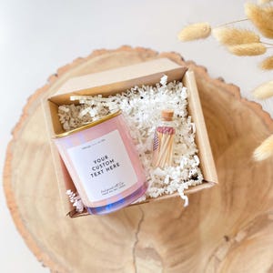 以下が含まれることがあります： 金色の蓋が付いたピンクのソイワックスキャンドルと、マッチが詰まった小さなガラス瓶が入ったギフトボックス。キャンドルにはラベルに「YOUR CUSTOM TEXT HERE」という文字が書かれています。箱は白いシュレッダーペーパーで満たされています。