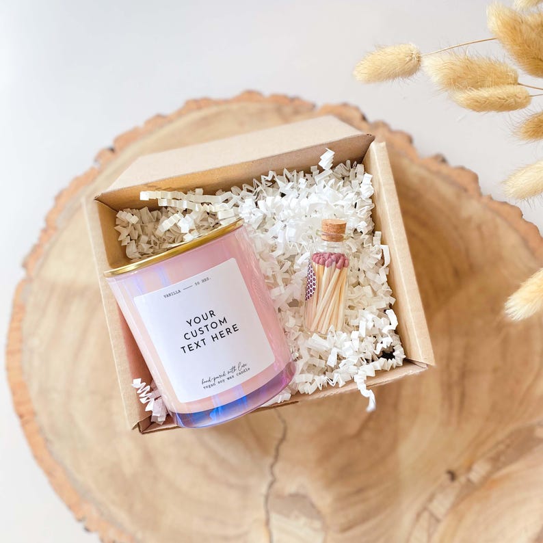 以下が含まれることがあります： 金色の蓋が付いたピンクのソイワックスキャンドルと、マッチが詰まった小さなガラス瓶が入ったギフトボックス。キャンドルにはラベルに「YOUR CUSTOM TEXT HERE」という文字が書かれています。箱は白いシュレッダーペーパーで満たされています。