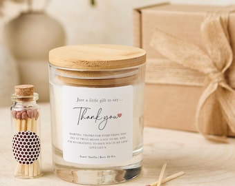 May include: A clear glass candle jar with a wooden lid and a label that says "Thank you." A small glass bottle of matches is next to the candle. A gift box with a ribbon is in the background. The candle scent is vanilla and has a 95-hour burn time.