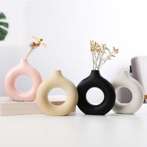 May include: Four ceramic vases in a row, each with a circular opening. The vases are in pastel pink, beige, black, and white. Each vase holds dried flowers. The vases are on a white surface.