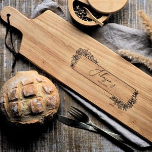 Puede incluir: Tabla de servir de madera con asa, grabada con el nombre "Johanson's" y detalles florales. La imagen incluye un pan, cubiertos y un cuenco pequeño con granos de pimienta.