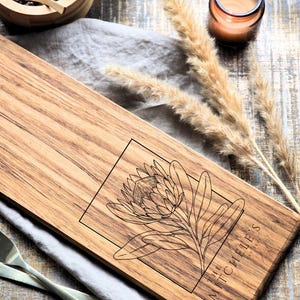 Puede incluir: Tabla de servir de madera con un diseño floral y el texto "THE MICHELE'S EST 2020". La tabla es rectangular y tiene vetas de madera natural. Se ve un cuenco de madera pequeño con granos de pimienta y una cuchara.