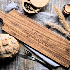 Puede incluir: Una tabla de servir de madera con asa, grabada con los nombres "Katie & Adam" y detalles florales. La tabla está junto a una hogaza de pan, cubiertos, un cuenco pequeño con granos de pimienta y una vela encendida.