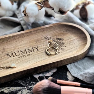 以下が含まれることがあります： 「Mummy We Love You」と刻まれた楕円形のウッドトレイ。トレイは薄い茶色で、金のリングが2つと白い石のネックレスが置かれている。