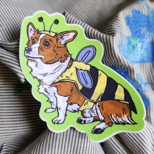 Può includere: Un adesivo con un cane corgi vestito da ape. Il corgi ha il pelo marrone e bianco, indossa un costume da ape a righe gialle e nere con ali blu e antenne. L'adesivo ha un bordo verde.