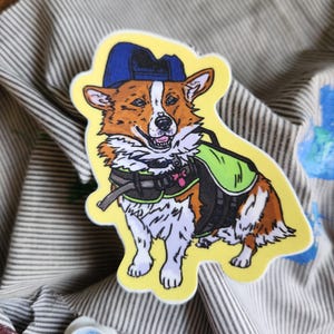 May include: A die-cut sticker featuring a smiling corgi wearing a blue cap, a green vest, and a black harness. The corgi has brown and white fur and is set against a yellow background.