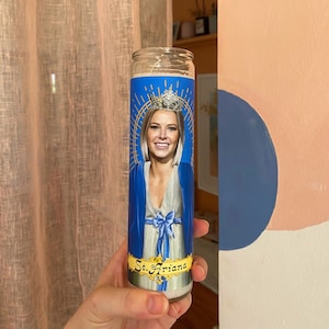Könnte beinhalten: Eine Glaskerze mit einem blauen und goldenen Etikett, auf dem eine Frau mit einer Krone und dem Text "St. Ariana" abgebildet ist.