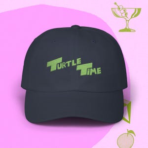 May include: A navy blue baseball cap with the words "TURTLE TIME" embroidered in green. The cap has a curved brim and a classic six-panel design. The background is a light pink color with green illustrations.