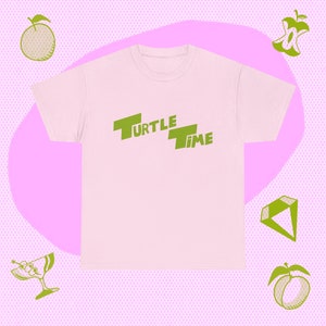 May include: A light pink t-shirt with the words "TURTLE TIME" printed in green. The shirt is surrounded by green illustrations of fruit and a cocktail glass on a pink and purple background.