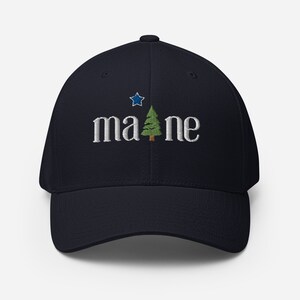 Può includere: Un cappello da baseball blu navy con un albero di pino bianco ricamato e la parola "maine" in lettere bianche. Una stella blu è sopra l'albero.
