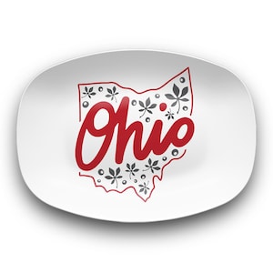 May include: A white, oval-shaped plate featuring a red outline of the state of Ohio. The state outline is filled with gray leaf and dot patterns. The word "Ohio" is written in red cursive script.