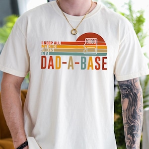 May include: Cream-colored t-shirt with the text "I KEEP ALL MY DAD JOKES IN A DAD-A-BASE" in a retro design. The graphic features a sunset-like image with a computer server. The shirt is short-sleeved and has a relaxed fit.