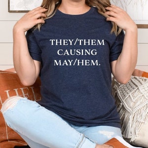 May include: A navy blue t-shirt with the text "THEY/THEM CAUSING MAY/HEM." printed in white.