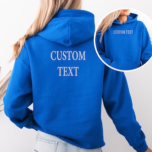May include: A bright blue hooded sweatshirt featuring the words CUSTOM TEXT printed in a light gray font on the back. An inset circular image shows the front of the same blue hoodie with the words CUSTOM TEXT printed on the upper chest area.