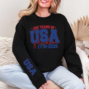 May include: Black sweatshirt with the text "250 YEARS OF USA freedom 1776-2026" in red and blue. The sleeve has the word "USA" in blue. The sweatshirt is a casual style.