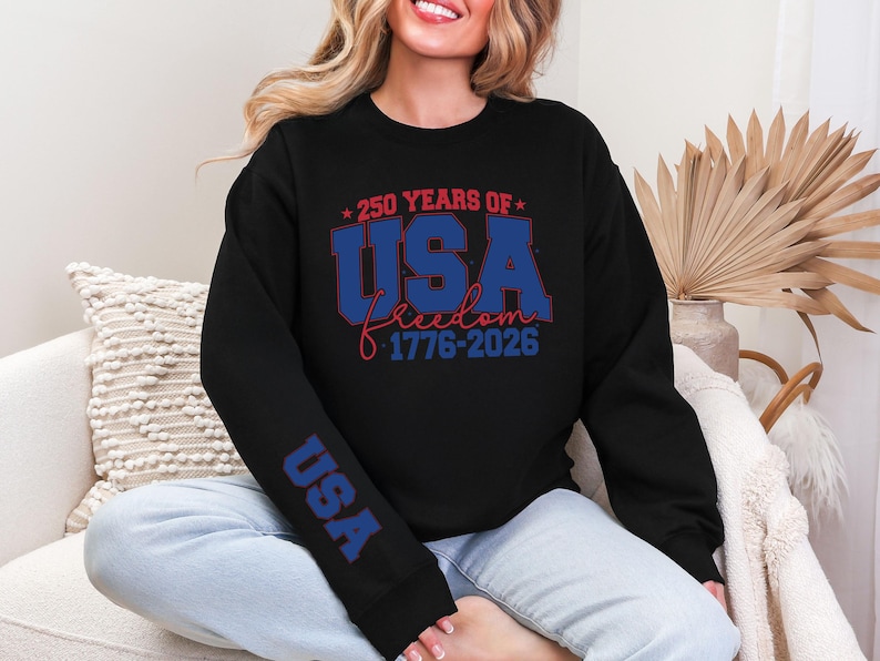 May include: Black sweatshirt with the text "250 YEARS OF USA freedom 1776-2026" in red and blue. The sleeve has the word "USA" in blue. The sweatshirt is a casual style.