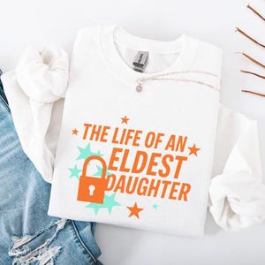 May include: White sweatshirt with the text "THE LIFE OF AN ELDEST DAUGHTER" in orange, with a lock graphic. The design includes orange stars and teal starbursts. A silver necklace is draped on the shirt.