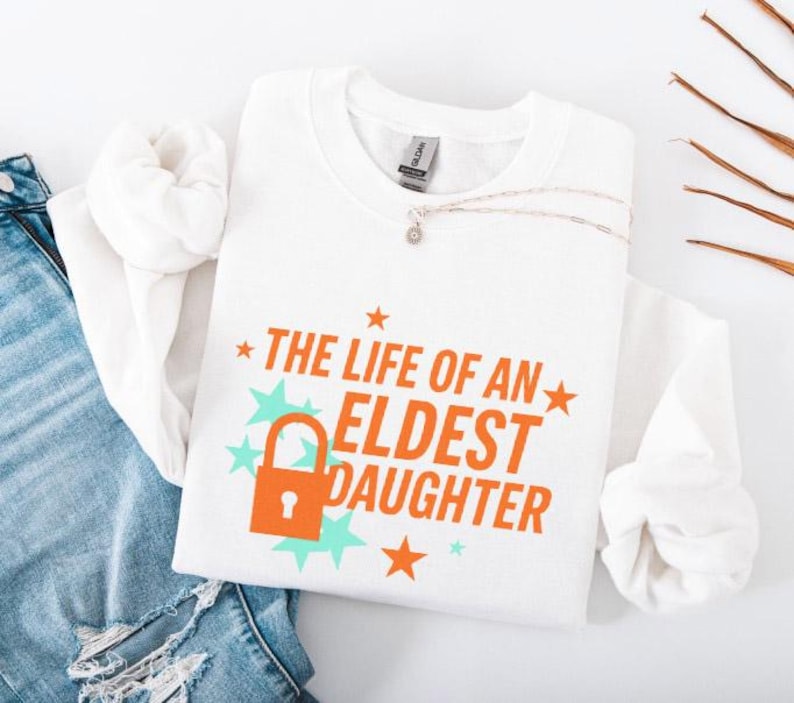 May include: White sweatshirt with the text "THE LIFE OF AN ELDEST DAUGHTER" in orange, with a lock graphic. The design includes orange stars and teal starbursts. A silver necklace is draped on the shirt.