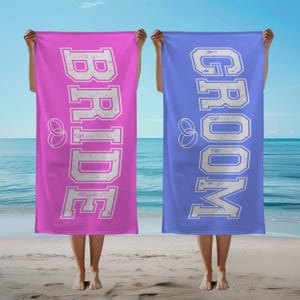 May include: Two beach towels, one pink and one blue, with the words "BRIDE" and "GROOM" in white, distressed lettering. Each towel has a white ring design.