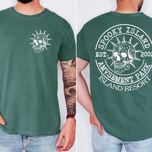 K&ouml;nnte beinhalten: Dunkelgr&uuml;nes T-Shirt mit wei&szlig;em Totenkopf-Design. Auf der Vorderseite befindet sich eine kleine Totenkopf-Grafik, w&auml;hrend die R&uuml;ckseite ein kreisf&ouml;rmiges Design mit dem Text "Spooky Island Amusement Park Island Resort" und dem Jahr "2002" zeigt.