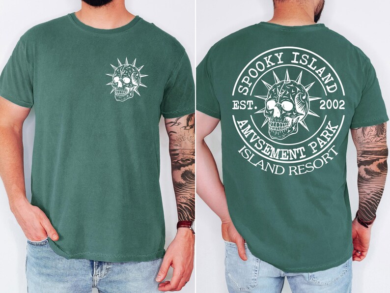 K&ouml;nnte beinhalten: Dunkelgr&uuml;nes T-Shirt mit wei&szlig;em Totenkopf-Design. Auf der Vorderseite befindet sich eine kleine Totenkopf-Grafik, w&auml;hrend die R&uuml;ckseite ein kreisf&ouml;rmiges Design mit dem Text "Spooky Island Amusement Park Island Resort" und dem Jahr "2002" zeigt.