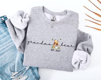 May include: A heather gray crewneck sweatshirt featuring the black cursive text "grandma bear" with a graphic of a cartoon bear holding a honey pot and the text "EST. 2027" below it. The sweatshirt is displayed alongside blue denim jeans.