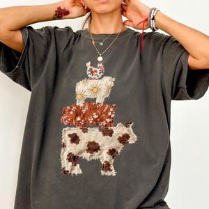 May include: Dark gray oversized t-shirt featuring a patchwork design of farm animals: a cow, a pig, and a chicken. The cow is brown and white, the pig is brown with a floral pattern, and the chicken is white with a floral pattern.