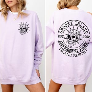 K&ouml;nnte beinhalten: Ein helllila Sweatshirt mit einem schwarzen Totenkopf-Motiv auf der Vorder- und R&uuml;ckseite. Das R&uuml;ckendesign ist ein kreisf&ouml;rmiges Design mit dem Text "Spooky Island Amusement Park Island Resort Est. 2002."