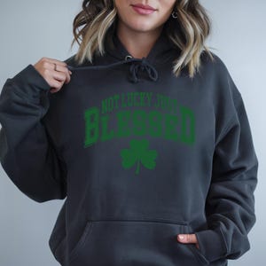 May include: A dark gray hoodie with a front pocket and a drawstring hood. The front of the hoodie features the text "NOT LUCKY JUST BLESSED" in green, arched over a green shamrock.