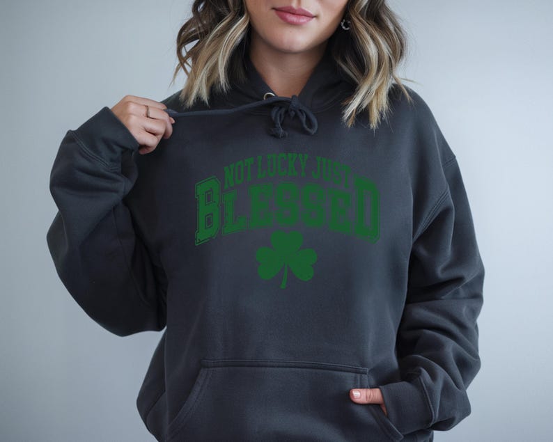 May include: A dark gray hoodie with a front pocket and a drawstring hood. The front of the hoodie features the text "NOT LUCKY JUST BLESSED" in green, arched over a green shamrock.