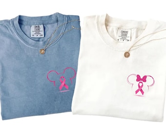 Könnte beinhalten: Zwei Comfort Colors T-Shirts, eines in Blau und eines in Weiß, jeweils mit einem rosa Mickey Mouse Kopf-Design und einem rosa Band, das das Bewusstsein für Brustkrebs symbolisiert. Goldene Halsketten mit runden Anhängern sind über die Shirts drapiert.
