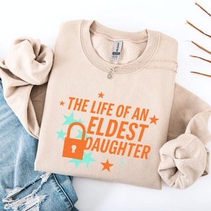 May include: A beige sweatshirt with the text "THE LIFE OF AN ELDEST DAUGHTER" in orange, with a lock graphic. The design includes teal stars and orange stars. A silver necklace is visible near the collar. The sweatshirt is folded on top of a pair of blue jeans.