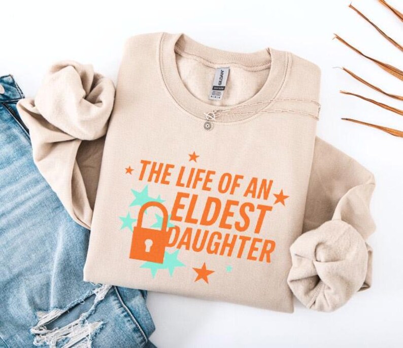 May include: A beige sweatshirt with the text "THE LIFE OF AN ELDEST DAUGHTER" in orange, with a lock graphic. The design includes teal stars and orange stars. A silver necklace is visible near the collar. The sweatshirt is folded on top of a pair of blue jeans.
