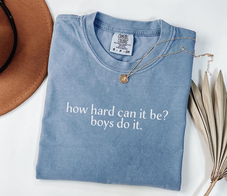 May include: A blue t-shirt with the text "how hard can it be? boys do it." A gold necklace with a pendant is on the shirt. The shirt is folded on a white surface.