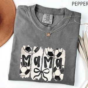 Puede incluir: Una camiseta gris Comfort Colors con un dise&ntilde;o de f&uacute;tbol. La palabra "Mama" est&aacute; impresa en negro, con balones de f&uacute;tbol a cada lado. Debajo de la palabra hay un lazo negro. Un collar dorado est&aacute; sobre la camiseta.