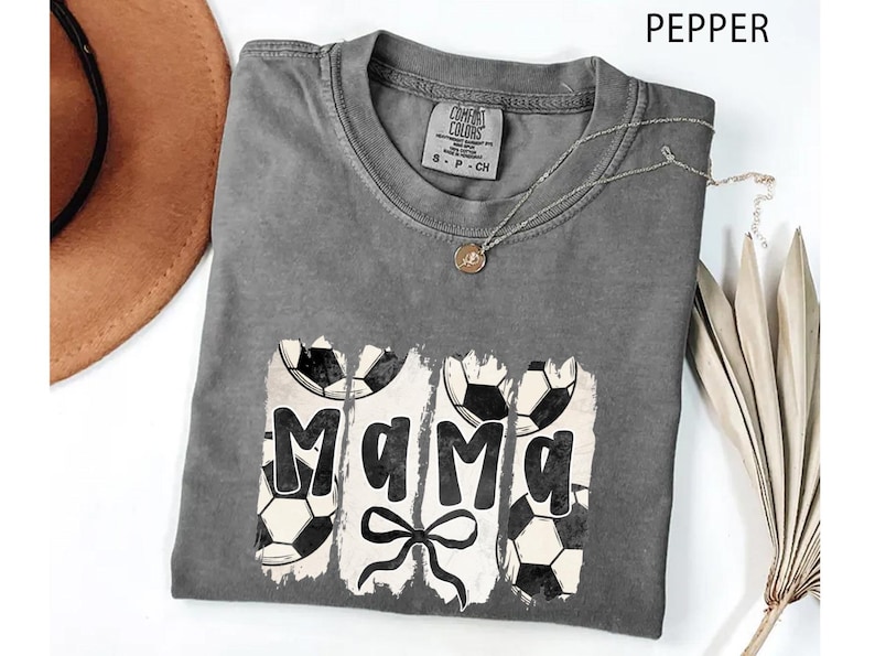 Puede incluir: Una camiseta gris Comfort Colors con un dise&ntilde;o de f&uacute;tbol. La palabra "Mama" est&aacute; impresa en negro, con balones de f&uacute;tbol a cada lado. Debajo de la palabra hay un lazo negro. Un collar dorado est&aacute; sobre la camiseta.