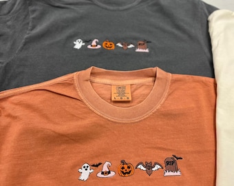 May include: A charcoal gray t-shirt and an orange t-shirt, both featuring an embroidered row of Halloween icons including a ghost, witch hat, pumpkin, bat, and tombstone with the text RIP. Each shirt has a Comfort Colors brand neck label.
