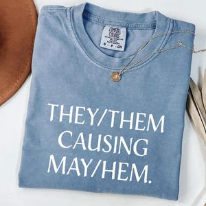 May include: A light blue t-shirt with the text "They/Them Causing May/Hem." printed in white.