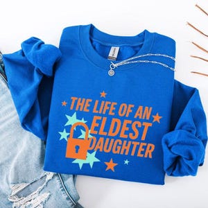 May include: A royal blue long-sleeve sweatshirt with the text "THE LIFE OF AN ELDEST DAUGHTER" in orange. The design includes a lock graphic and star shapes. A silver necklace with a pendant is on the shirt.
