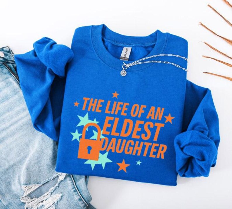 May include: A royal blue long-sleeve sweatshirt with the text "THE LIFE OF AN ELDEST DAUGHTER" in orange. The design includes a lock graphic and star shapes. A silver necklace with a pendant is on the shirt.