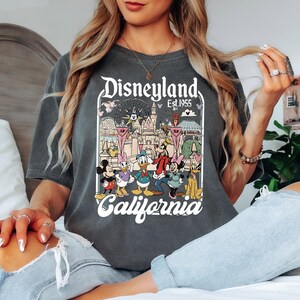May include: Dark gray t-shirt featuring a Disneyland graphic with Mickey Mouse, Minnie Mouse, Donald Duck, Goofy, and other characters. The graphic includes the text "Disneyland Est. 1955 California". The shirt has a vintage, distressed look.
