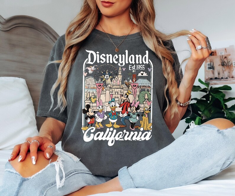 May include: Dark gray t-shirt featuring a Disneyland graphic with Mickey Mouse, Minnie Mouse, Donald Duck, Goofy, and other characters. The graphic includes the text "Disneyland Est. 1955 California". The shirt has a vintage, distressed look.