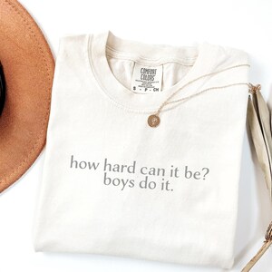 May include: Cream-coloured t-shirt with the text "how hard can it be? boys do it." A gold necklace with a pendant rests on the shirt. A brown hat and dried plant are in the background.
