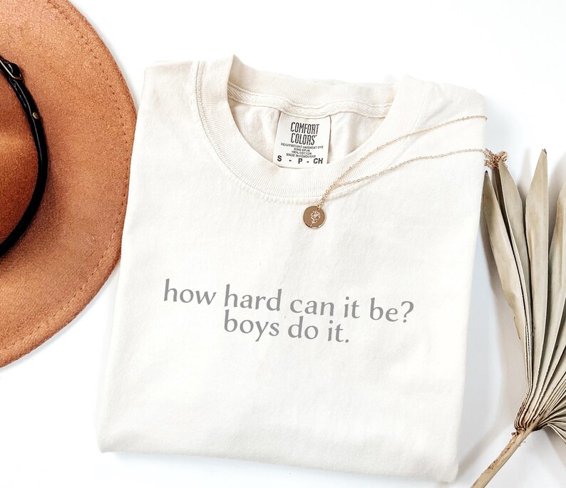 May include: Cream-coloured t-shirt with the text "how hard can it be? boys do it." A gold necklace with a pendant rests on the shirt. A brown hat and dried plant are in the background.